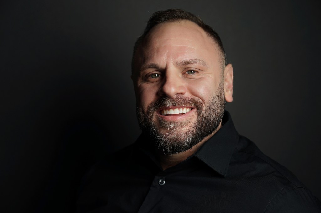 Professional headshot of a bearded man smiling wearing a black shirt with a black background in Phoenix Arizona